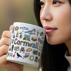 Op de afbeelding: Witte keramische mok met het woord "Cornwall" en diverse kleurrijke illustraties van lokale bezienswaardigheden en symbolen. De mok toont afbeeldingen van een zeehond, vis en ijs.