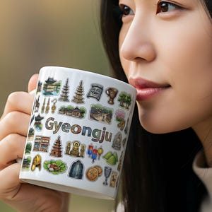 May include: White ceramic mug with illustrations of Gyeongju landmarks. The mug has a handle and is decorated with various images and the city's name. A person is holding the mug.