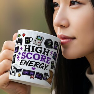 May include: White ceramic mug with a colourful design featuring video game-themed graphics and the words "HIGH SCORE ENERGY." The mug includes icons of game controllers, a handheld console, and the text "GAME ON."
