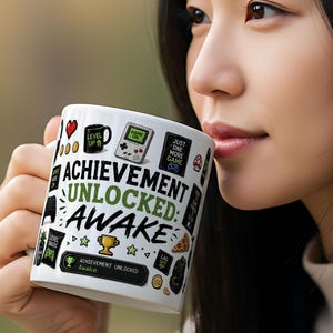 Peut inclure: Tasse en céramique blanche avec l'inscription "ACHIEVEMENT UNLOCKED: AWAKE" et divers graphismes sur le thème des jeux vidéo. La tasse présente des icônes telles qu'un cœur, une tasse à café, une console de jeu et un trophée. La tasse est tenue par une personne.