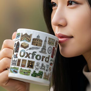 Op de afbeelding: Witte keramische mok met het woord "Oxford" en diverse Oxford-bezienswaardigheden. De mok is versierd met kleurrijke illustraties van gebouwen, monumenten en andere symbolen. De mok wordt door een persoon vastgehouden.