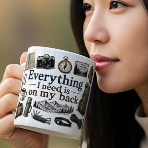 May include: White ceramic mug featuring the text "Everything I need is on my back" and illustrations of outdoor equipment, including a camera and compass. The mug is held by a person.