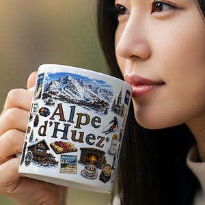 Pode incluir: Caneca de cerâmica branca com o texto "Alpe d'Huez" e ilustrações de uma cordilheira nevada, um esquiador e um chalé. A caneca é segurada por uma pessoa.