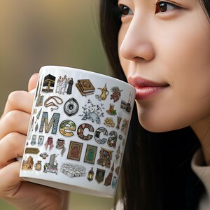 Könnte beinhalten: Weißer Keramikbecher mit bunten Illustrationen und dem Wort "Mecca". Der Becher ist mit verschiedenen religiösen Symbolen und Bildern verziert. Der Becher wird von einer Person gehalten.