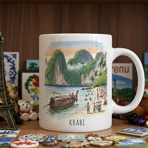 May include: White ceramic mug with a scenic illustration of Krabi, Thailand, featuring a longtail boat, beach, and mountains. The word "KRABI" is printed at the bottom. The mug has a curved handle.