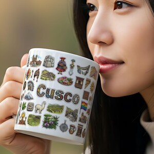 Może przedstawiać: Biały ceramiczny kubek z kolorowymi ilustracjami Cusco w Peru i nazwą miasta. Kubek jest trzymany przez osobę, z naciskiem na szczegółowy design i słowo "Cusco".