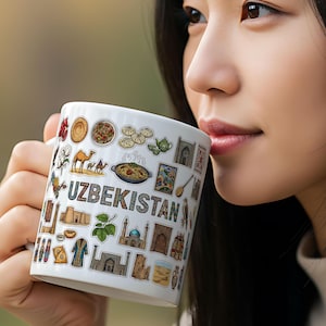 May include: White ceramic mug featuring the word "UZBEKISTAN" and various colorful illustrations of Uzbek culture, including food, architecture, and camels. The mug is held by a person.
