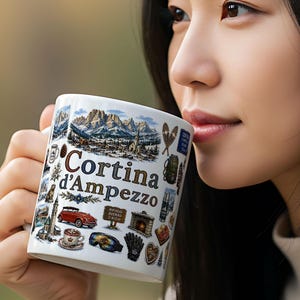 Może przedstawiać: Biały ceramiczny kubek z napisem "Cortina d'Ampezzo" i ilustracjami łańcucha górskiego, czerwonego samochodu i innych ikon. Kubek ma uchwyt i jest wypełniony napojem.