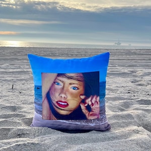 May include: A blue throw pillow with a square image of a woman's face with blue eyes and red lips. The image is set against a blue and white ocean background.