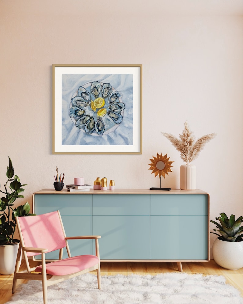 May include: A watercolor painting of a plate of oysters with lemon wedges. The oysters are arranged in a circle on a white plate with a blue background. The painting is framed in a light wood frame and hangs on a light pink wall.