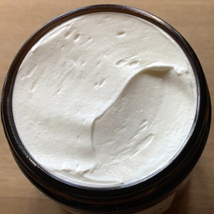 Puede incluir: Una vista cercana de una crema corporal blanca y cremosa en un frasco de vidrio con una tapa marr&oacute;n oscura. La crema es suave y tiene un patr&oacute;n de remolino.
