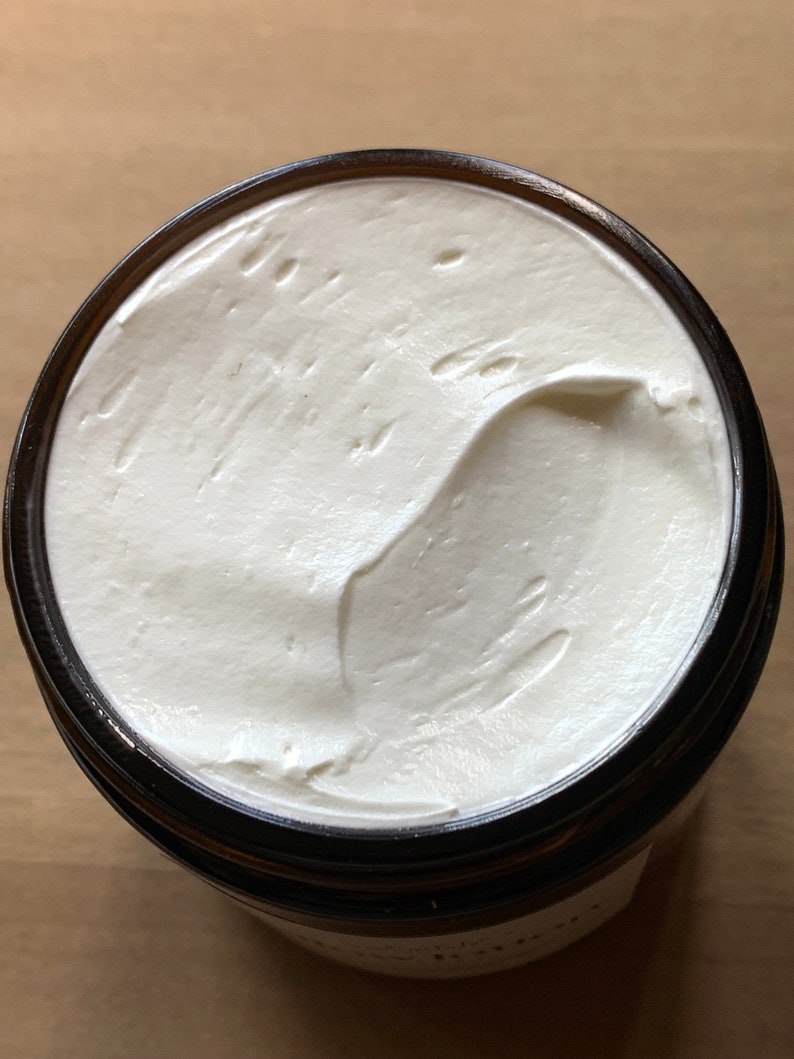 Puede incluir: Una vista cercana de una crema corporal blanca y cremosa en un frasco de vidrio con una tapa marr&oacute;n oscura. La crema es suave y tiene un patr&oacute;n de remolino.