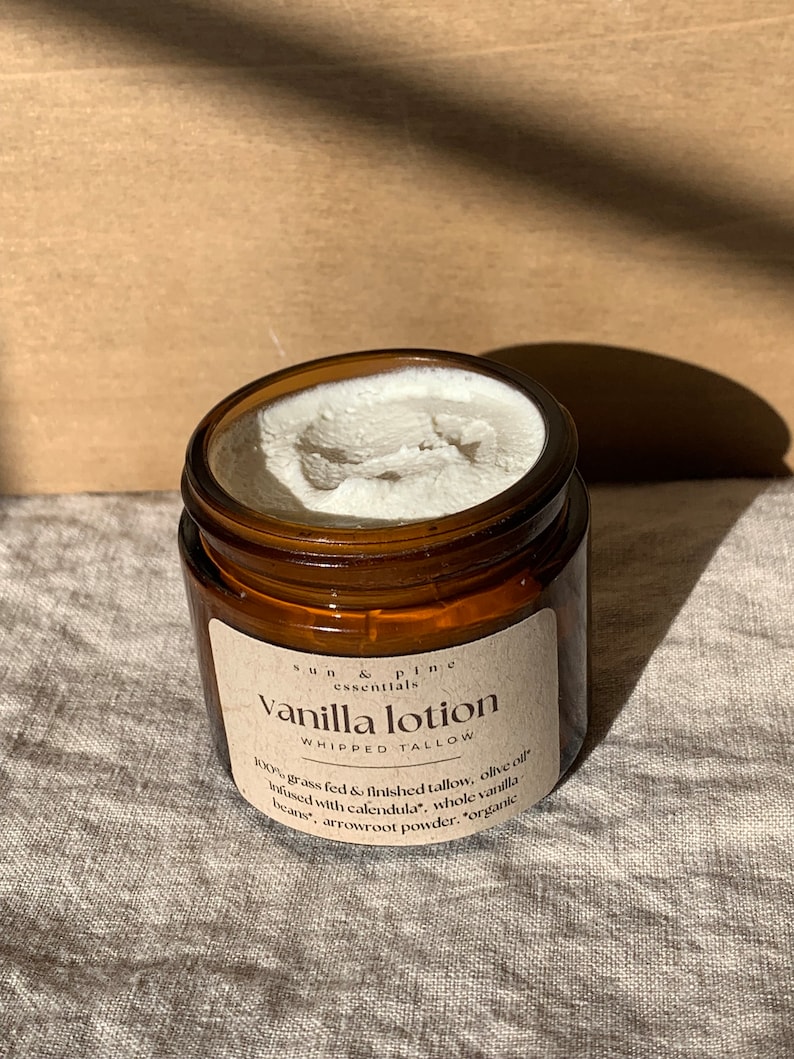 Puede incluir: Un tarro de vidrio marr&oacute;n con una etiqueta blanca que dice "Sun & Pine Essentials Vanilla Lotion Whipped Tallow 100% grass fed & finished tallow, olive oil infused with calendula, whole vanilla beans, arrowroot powder, organic"