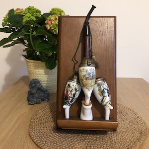 May include: A set of three porcelain pipes with painted designs, displayed on a wooden stand with a long pipe hanging above. The pipes are decorated with colorful scenes and have a vintage aesthetic.