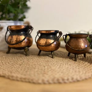 May include: Three small copper cauldrons with black accents and handles. The cauldrons are sitting on a woven brown mat.