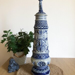 Op de afbeelding: Een hoge, blauw-witte keramische steen met ingewikkelde ontwerpen en een beeldje bovenop. De steen heeft een blauw-witte banner met de tekst "Trink  mut".