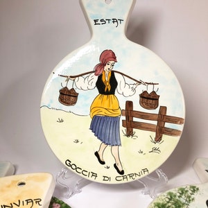 May include: A white ceramic trivet with a hand-painted design of a woman carrying two baskets on a stick. The woman is wearing a yellow skirt, blue blouse, and red headscarf. The trivet has the words "ESTAT GOCCIA DI CARNIA" written on it.