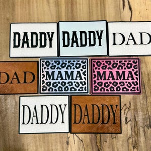 May include: A collection of rectangular patches with the words "DADDY", "DAD", and "MAMA" in bold, black lettering. Some patches have a textured background, while others feature leopard print patterns in various colors, including pink and blue.