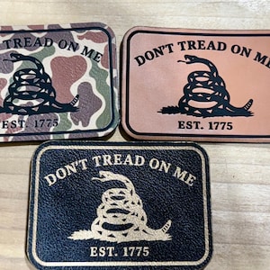 May include: Three rectangular patches with the text "DON'T TREAD ON ME" and "EST. 1775". One patch has a camouflage pattern, another is tan, and the third is navy blue. Each features a coiled snake.