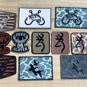 May include: A collection of patches featuring hunting and fishing themes. Designs include deer, fish on hooks, and waterfowl, with some incorporating camouflage patterns and the American flag. Patches vary in shape and color.