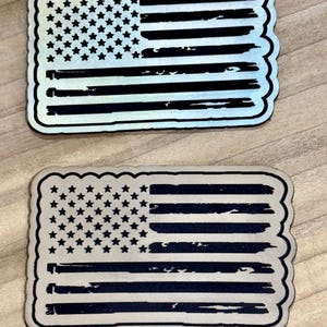 May include: Two rectangular stickers featuring the American flag design. The flags have a distressed look with black stripes and a field of stars. The stickers have a scalloped edge and are set against a light wood grain background.