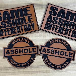 May include: Four patches with humorous text. Two rectangular patches read "SAME ASSHOLE DIFFERENT DAY" in black letters on a brown background. Two circular patches read "GENUINE ASSHOLE MADE IN THE U.S.A." in black and brown.