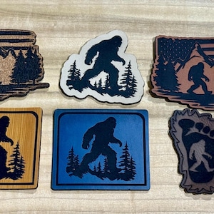 May include: A collection of Bigfoot-themed wooden and acrylic cutouts. Designs include the silhouette of Bigfoot walking through a forest, with some featuring an American flag and mountain scene. Various shapes and colors.