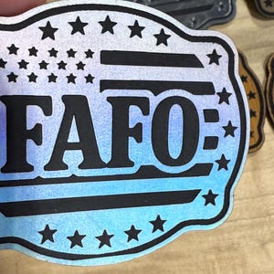 May include: A wooden badge with a design resembling the American flag, featuring the text "FAFO" in bold black letters. The badge has a blue and white gradient background and is surrounded by stars.