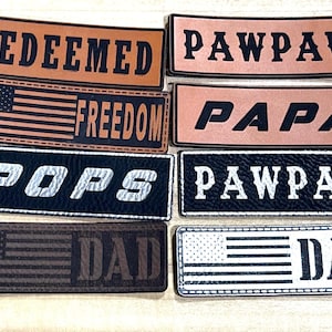 May include: A collection of rectangular patches in various colors, featuring text and American flag designs. The patches display words like "REDEEMED," "FREEDOM," "POPS," "DAD," and "PAWPAW," in bold, contrasting fonts. The patches are likely for clothing or accessories.