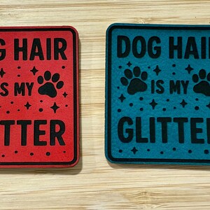 May include: Two square signs with the text "DOG HAIR IS MY GLITTER". One sign is red with black text and paw prints, the other is teal with black text and paw prints. Both have a black border.