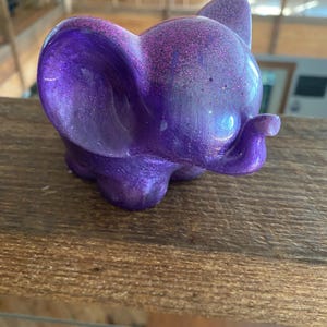 Może przedstawiać: Mała, fioletowa figurka słonia z brokatem osadzonym w żywicy. Figurka ma gładkie, błyszczące wykończenie.