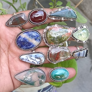 May include: A collection of nine silver rings featuring a variety of gemstone settings. The rings showcase a range of colours and patterns, including green, blue, red, and white. The stones are set in silver bezels and are surrounded by decorative beading.