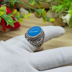May include: A silver ring with an oval, light blue stone. The ring has an ornate, detailed design around the stone and on the band. The ring is displayed on a white surface.