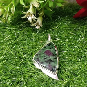 May include: A silver pendant with a triangular, polished stone. The stone is a mix of dark green and black with a prominent, round, deep red inclusion. The pendant is set against a backdrop of green grass.