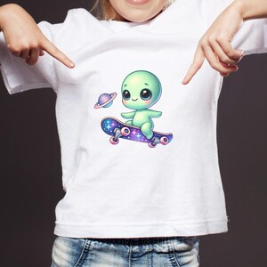Puede incluir: Una camiseta blanca con un extraterrestre verde haciendo skateboard en una tabla de skate con tem&aacute;tica de galaxia p&uacute;rpura y azul. El extraterrestre sostiene un peque&ntilde;o planeta en su mano.
