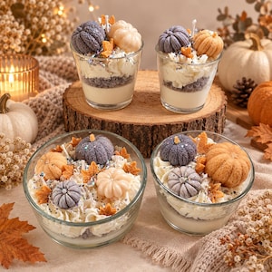 Puede incluir: Cuatro recipientes de vidrio rellenos de cera blanca, cubiertos con cera blanca batida, pequeñas figuras de cera con forma de calabaza en tonos morado, naranja y crema, y hojas de cera naranja esparcidas con purpurina iridiscente.