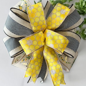 May include: A yellow and black bow with a honeycomb pattern and a bee design. The bow is made of ribbon and is tied in a knot.
