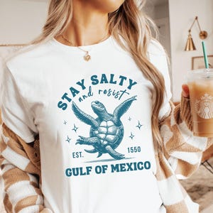 Peut inclure: T-shirt blanc avec un motif bleu d'une tortue de mer et le texte "Stay Salty and Resist". Le t-shirt comprend également le texte "Gulf of Mexico" et le chiffre "1550."