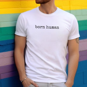 May include: White t-shirt with the text "born human" in a black, simple font. The shirt is plain and has short sleeves. The background is a colorful wall with horizontal stripes of yellow, orange, blue, and purple.
