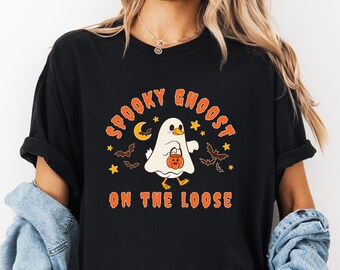 May include: Black t-shirt with a Halloween-themed graphic. The design features a white ghost with a pumpkin bucket, a crescent moon, stars, and bats. The text reads "Spooky Ghoost On The Loose" in orange.