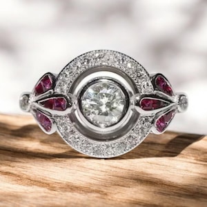 May include: A silver ring with a round, clear center stone surrounded by smaller diamonds. The ring has two wing-shaped accents on either side of the center stone, set with red rubies.