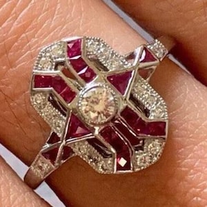May include: A silver ring with a large, geometric design featuring a central diamond surrounded by ruby stones.