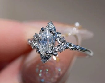 May include: A silver ring with a marquise-shaped diamond set in a delicate, ornate design. The ring features intricate details and smaller diamonds surrounding the center stone.