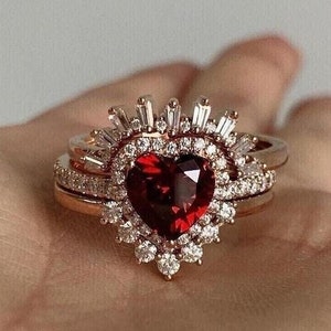 May include: A heart-shaped ruby engagement ring with a halo of smaller diamonds set in a rose gold band. The ring is surrounded by a delicate band of smaller diamonds.