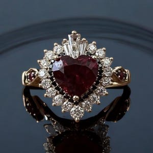Puede incluir: Un anillo de oro con un gran rubí en forma de corazón rodeado por un halo de diamantes más pequeños y diamantes negros.