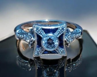 May include: A silver ring with a square, dark blue center stone and a round, light blue center stone. The ring has intricate detailing with smaller, clear stones and is set against a reflective surface.
