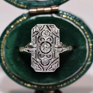 Op de afbeelding: Een sierlijke ring in Art Deco-stijl met een rechthoekige vorm, bezet met een centrale diamant en kleinere stenen. De ring wordt gepresenteerd in een groene fluwelen ringdoos met een gouden rand, wat een luxueuze presentatie creëert.