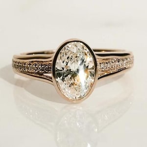 Könnte beinhalten: Ein Verlobungsring aus Roségold mit einem großen ovalen Diamanten, der in einer Zargenfassung sitzt. Die Ringschiene ist mit kleineren Diamanten besetzt.