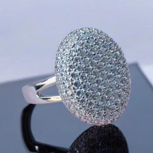 Puede incluir: Un anillo de plata con una gran piedra central de forma ovalada. La piedra está cubierta de pequeños circonios cúbicos brillantes.