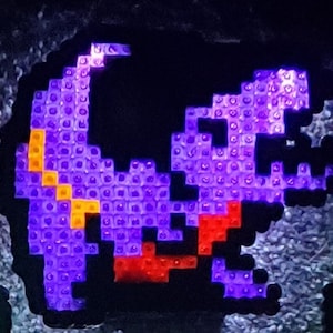 May include: A purple and red pixelated dinosaur made of small plastic squares. The dinosaur is facing right and has a long tail.